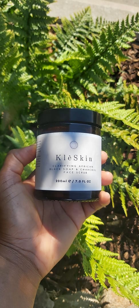 a person holding an jar of Clarifying African Black Soap & Charcoal Face Scrub with green fern plant in the background