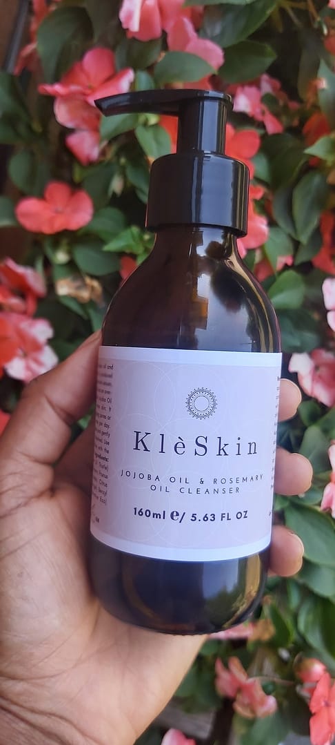 a person's hand holding a bottle of jojoba & rosemary face oil cleanser with flowers in the background