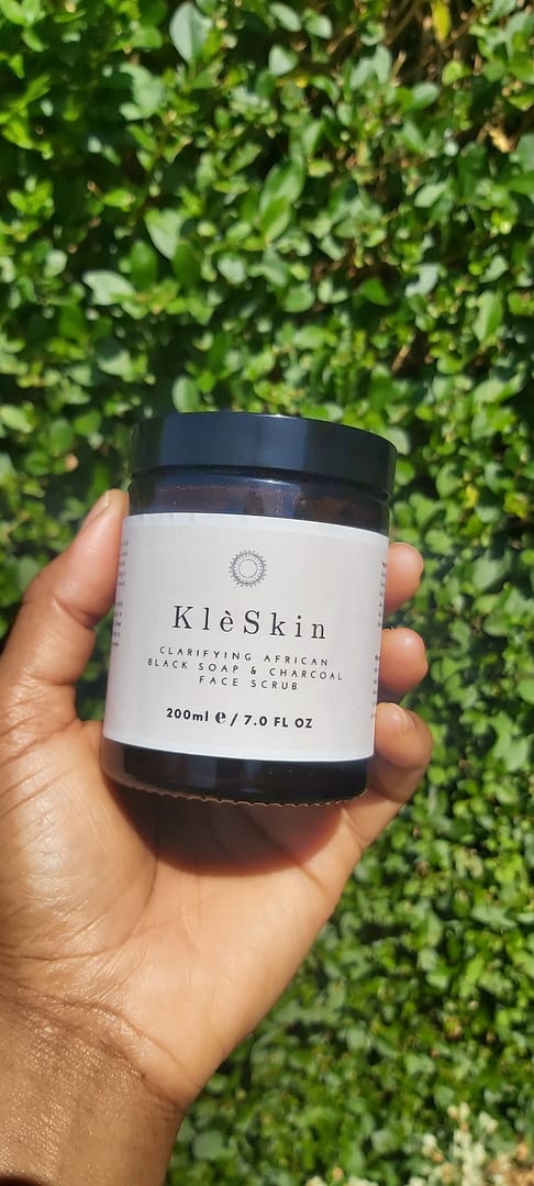 a person holding a jar of Clarifying African Black Soap & Charcoal Face Scrub with green hedge in the background