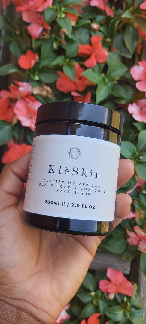 a person holding a jar of Clarifying African Black Soap & Charcoal Face Scrub with pink and green flowers in the background