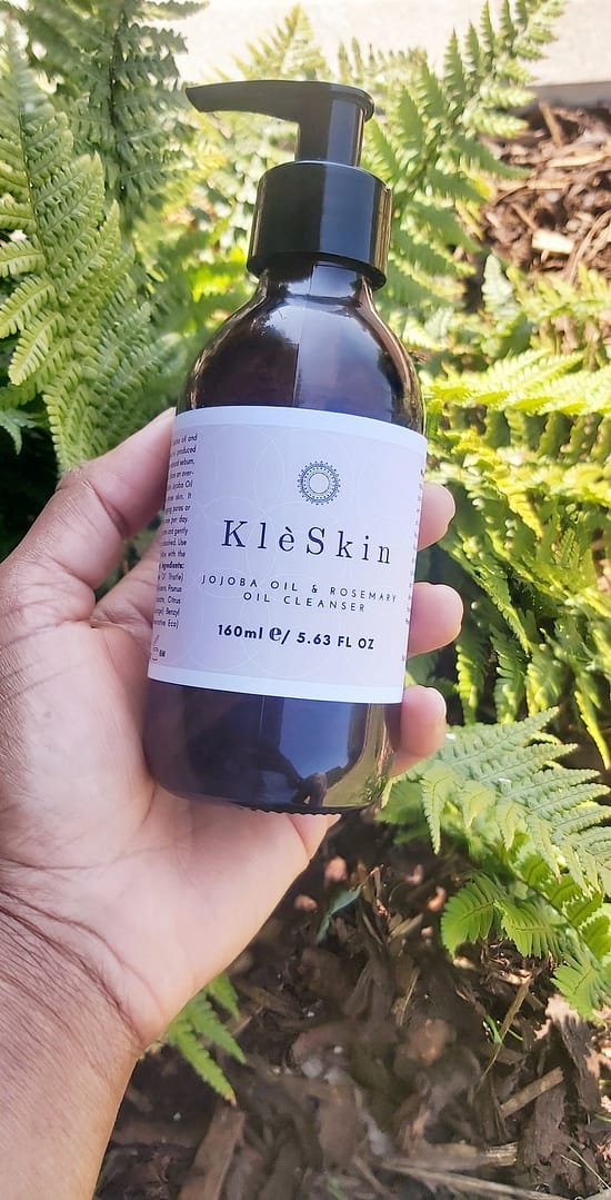 a person's hand holding a bottle of jojoba & rosemary face oil cleanser with green fern in the background